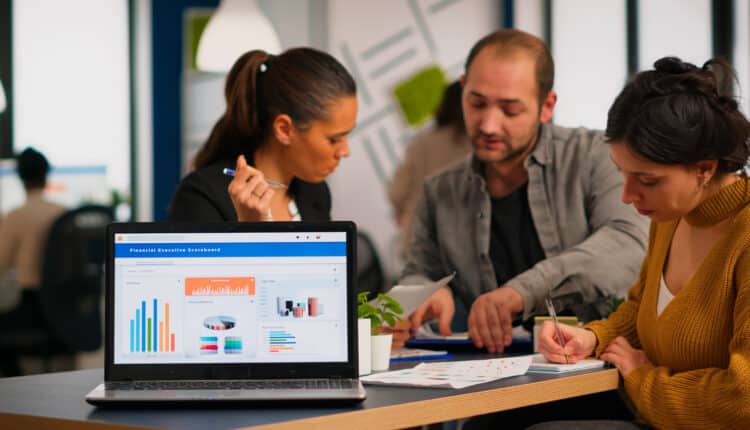 Businesswoman discussing a financial project while taking notes laptop, in a co-working space with diverse team members collaborating.