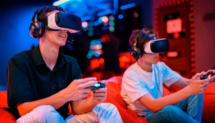 Two teenage friends at home, one wearing a VR headset and headphones and both holding game controllers, immersed in a console video game.